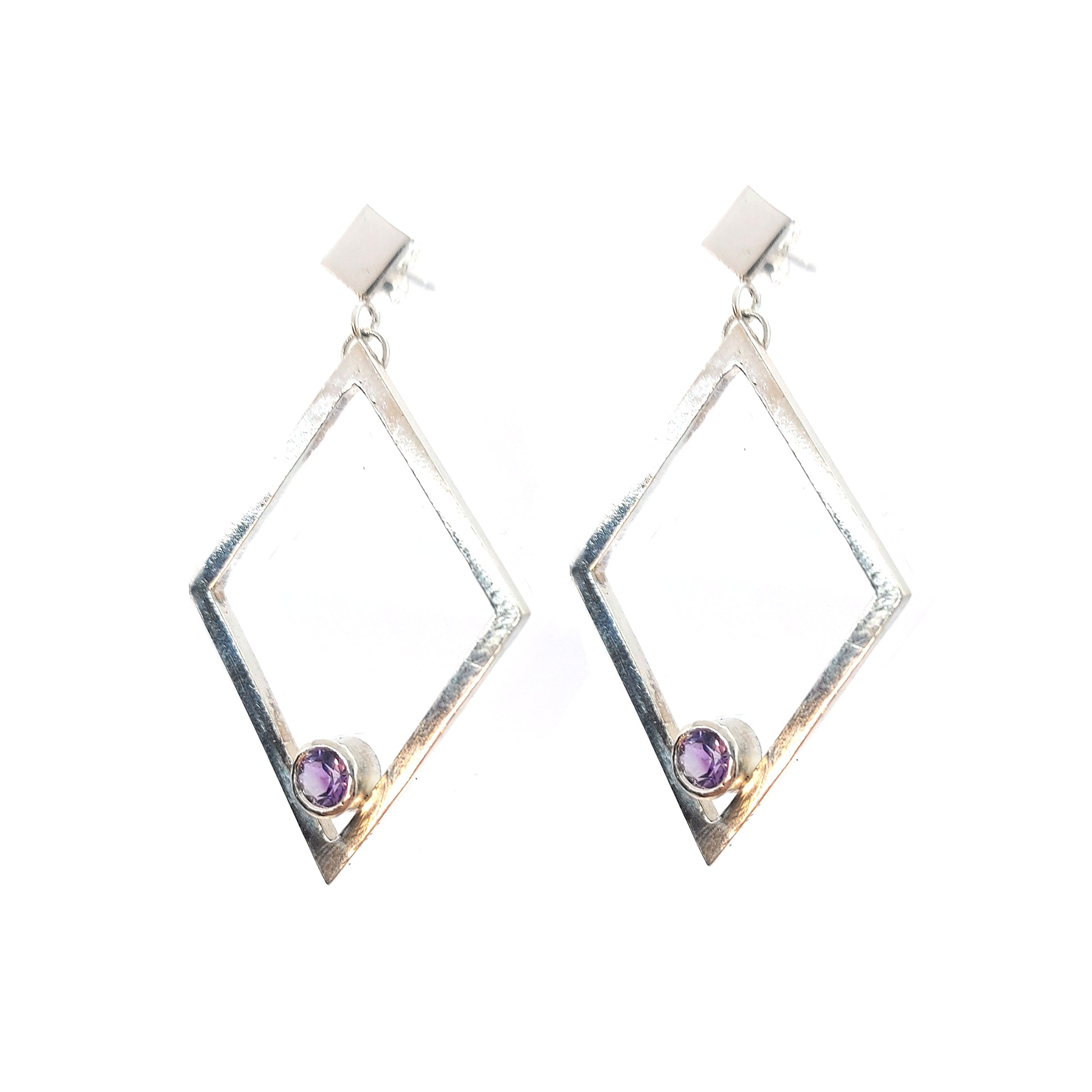 A pair of silver statement earrings featuring a top diamond shaped stud with a large open diamond shape drop beneath. Inside at the bottom of the large drop diamond is a round purple amethyst.