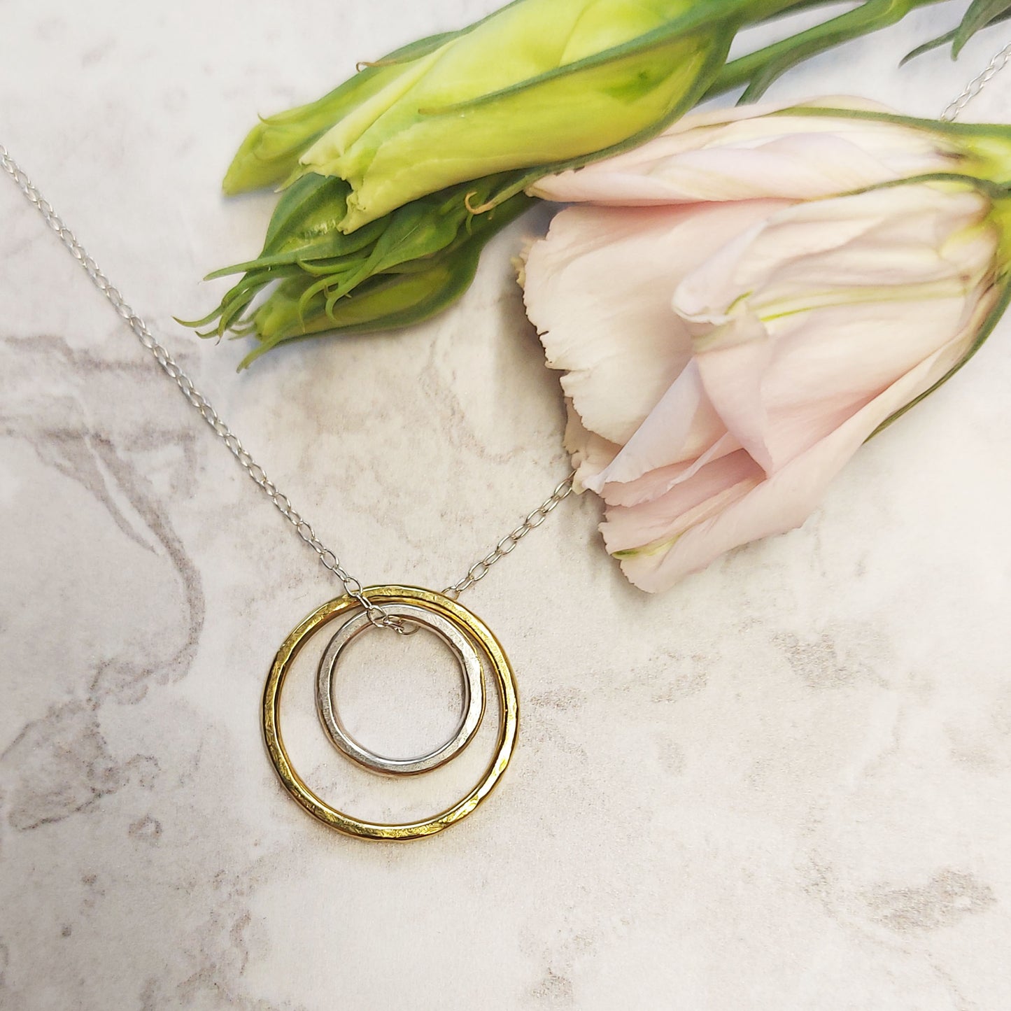 A double circle pendant with an inner silver hammered circle and an outer yellow gold vermeil hammered circle. Suspended on a silver chain. Pictured with flowers.