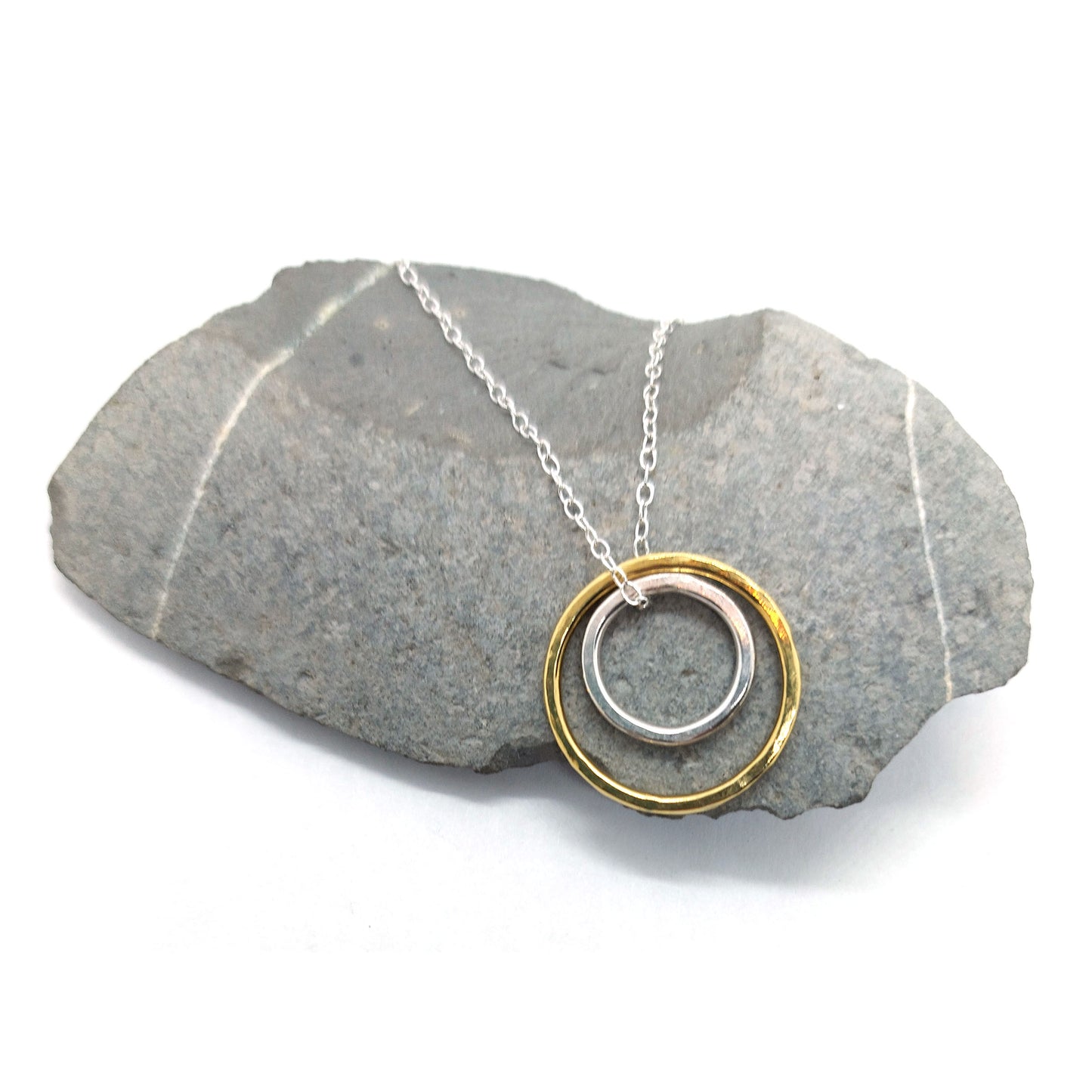 A double circle pendant with an inner silver hammered circle and an outer yellow gold vermeil hammered circle. Suspended on a silver chain. Pictured on a pebble.