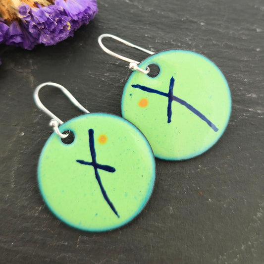 A pair of handmade round green enamel drop earrings with an asymmetric blue cross and orange dot. On sterling silver ear hooks. On a black background with purple flowers. Mijoux jewellery.
