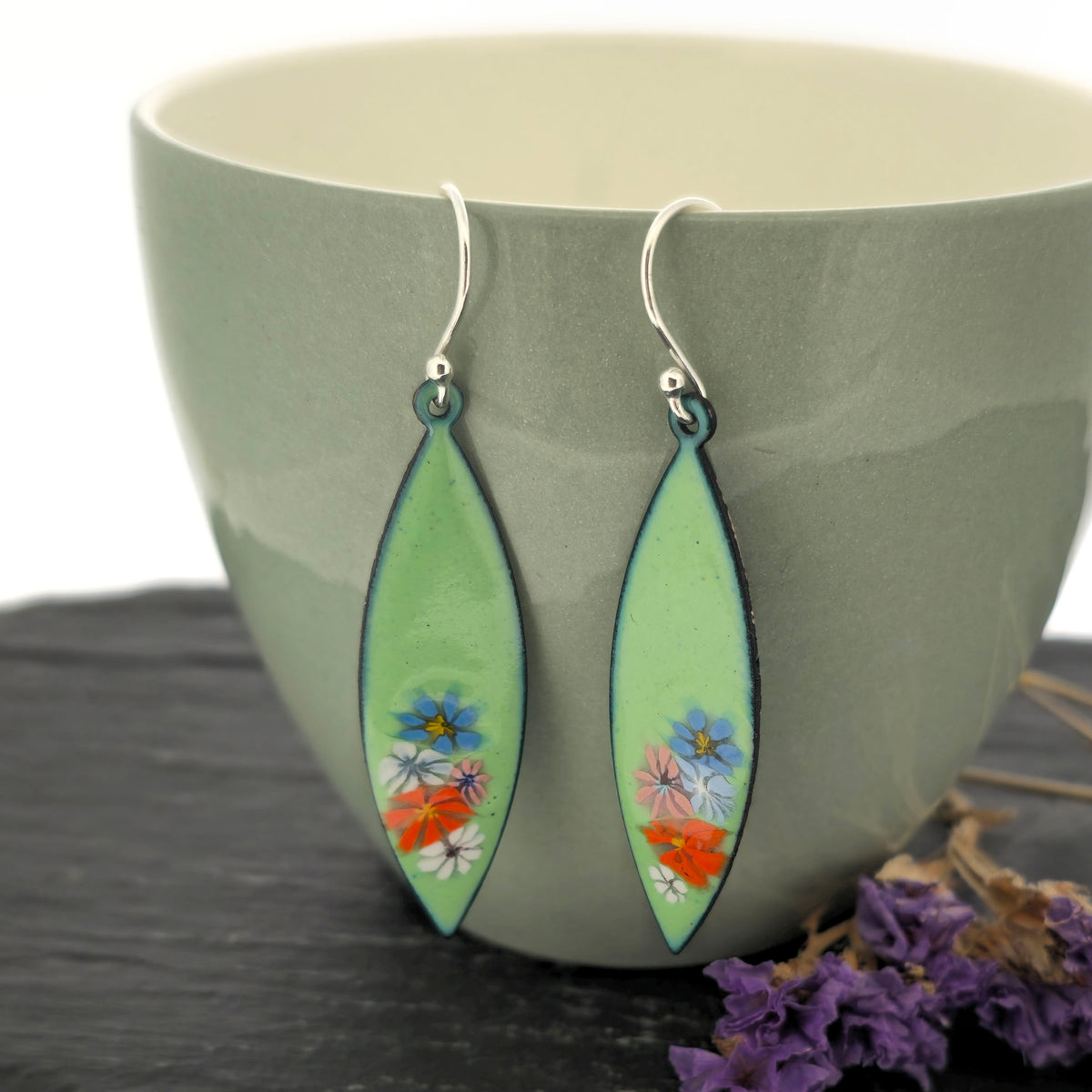 A pair of dangling earrings with elongated light green enamel leaf-shaped drops containing enamel flowers, hanging from silver hooks on a sage green cup. Mijoux jewellery.