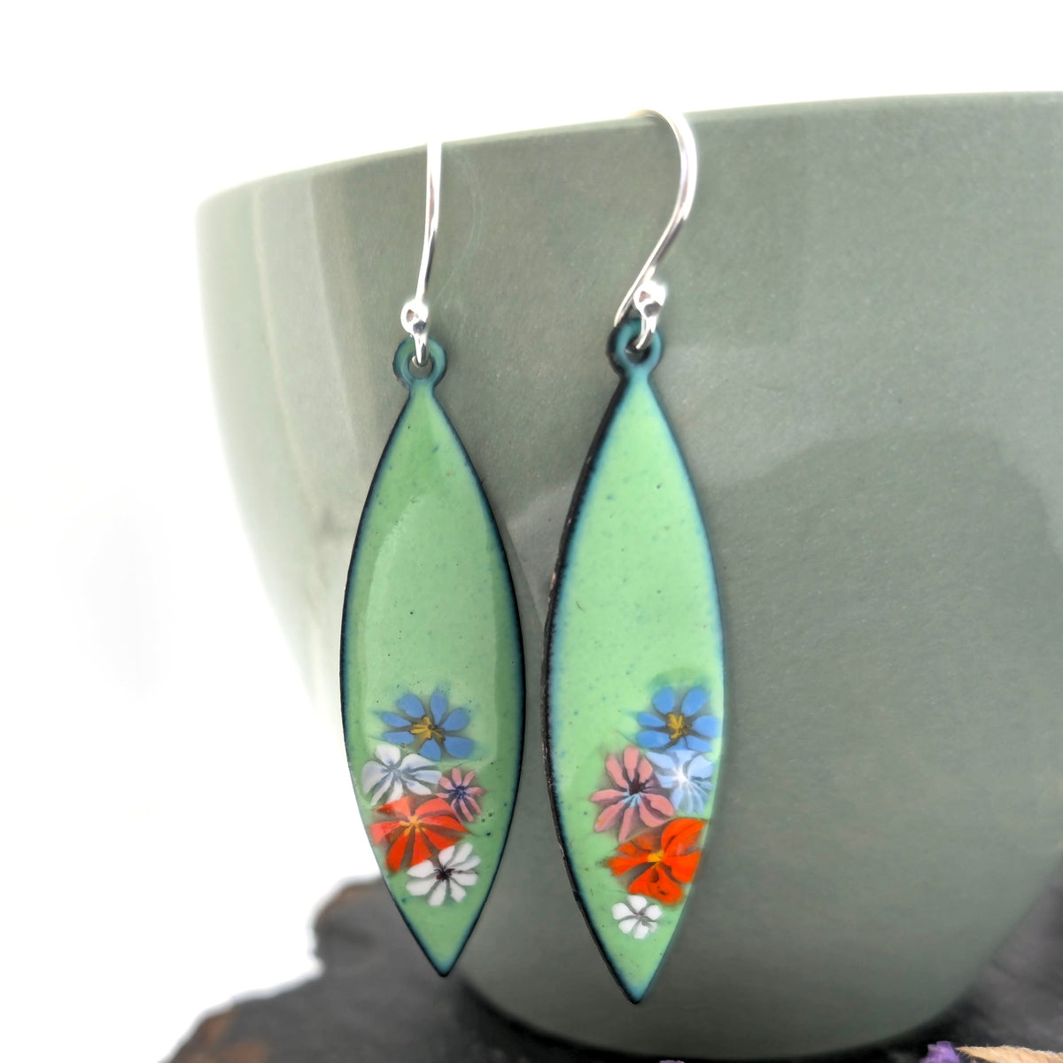 A pair of handmade dangling marquise-shaped earrings with a light green background and small colorful enamel floral details, hanging in front of a muted green ceramic cup. Mijoux jewellery.