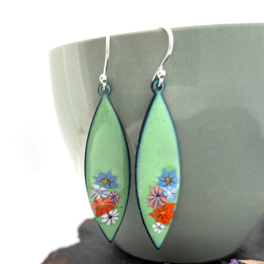 A pair of handmade dangling marquise-shaped earrings with a light green background and small colorful enamel floral details, hanging in front of a muted green ceramic cup. Mijoux jewellery.