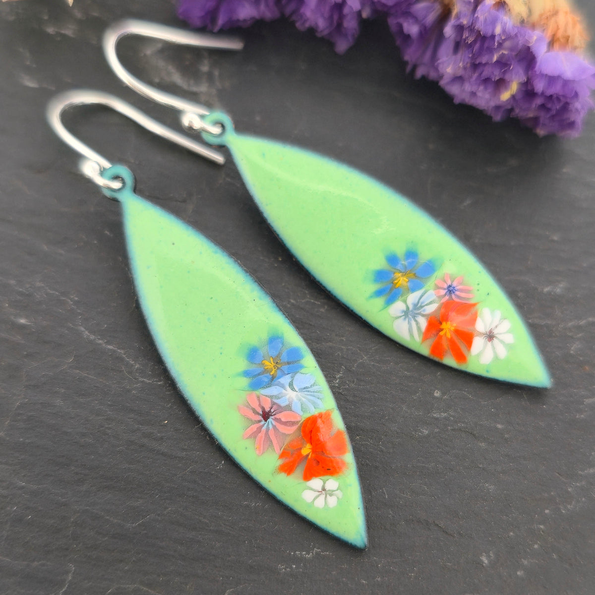 A pair of handmade dangling marquise-shaped earrings with a light green background and small colorful enamel floral details, on a black background. Mijoux jewellery.