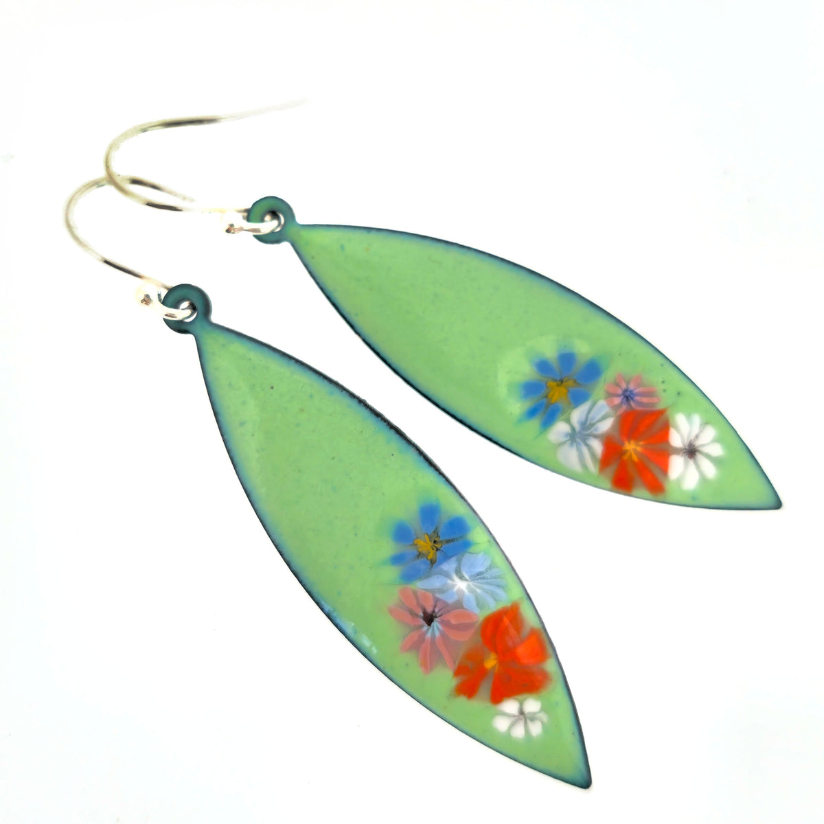 A pair of dangling earrings with elongated light green enamel leaf-shaped drops containing enamel flowers, hanging from silver hooks against a plain white background. Mijoux jewellery.