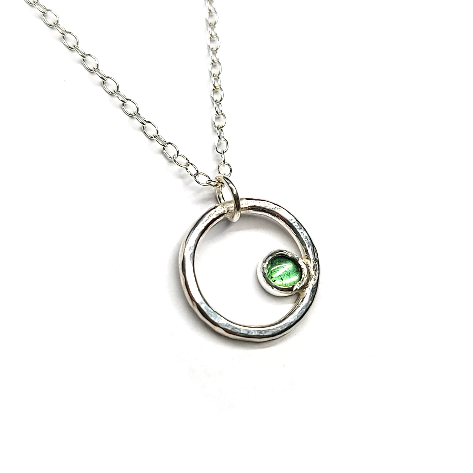 Silver hammered open circle pendant with round green tourmaline gemstone set off-centre. On a silver chain.