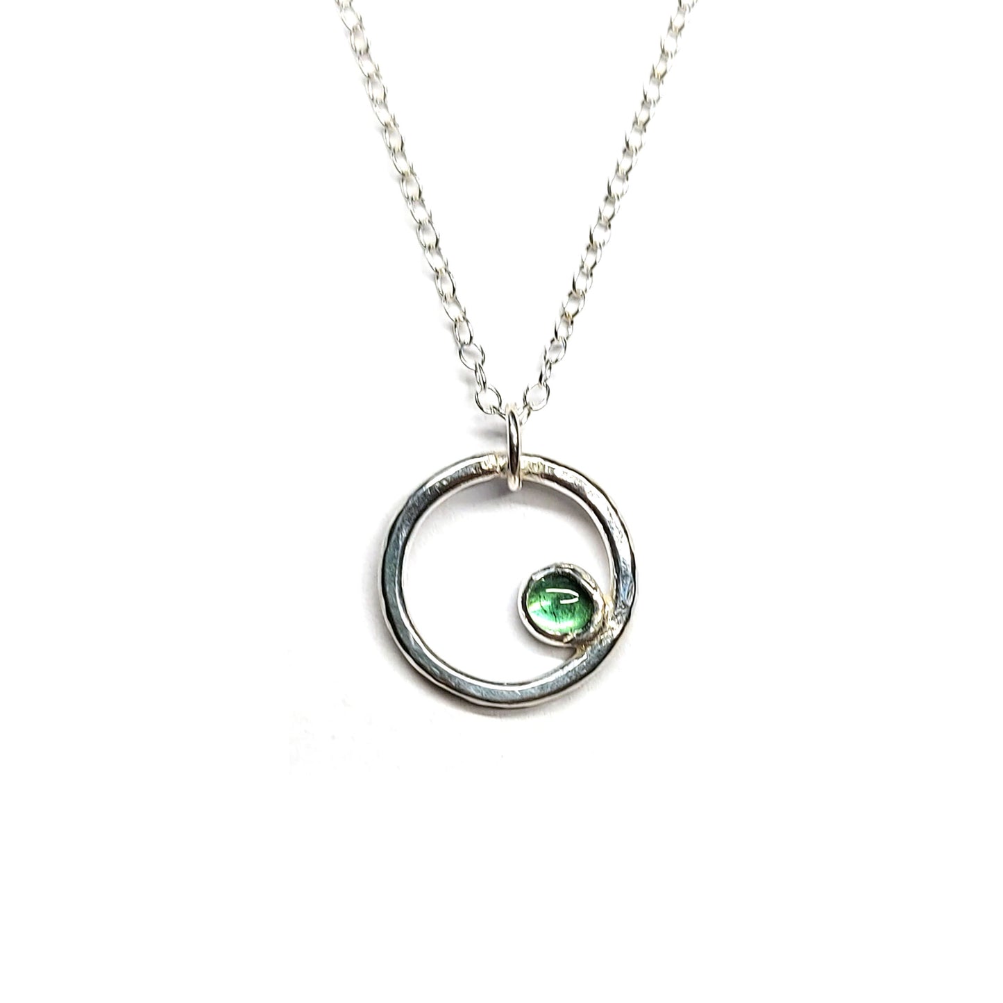 Silver hammered open circle pendant with round green tourmaline gemstone set off-centre. On a silver chain.