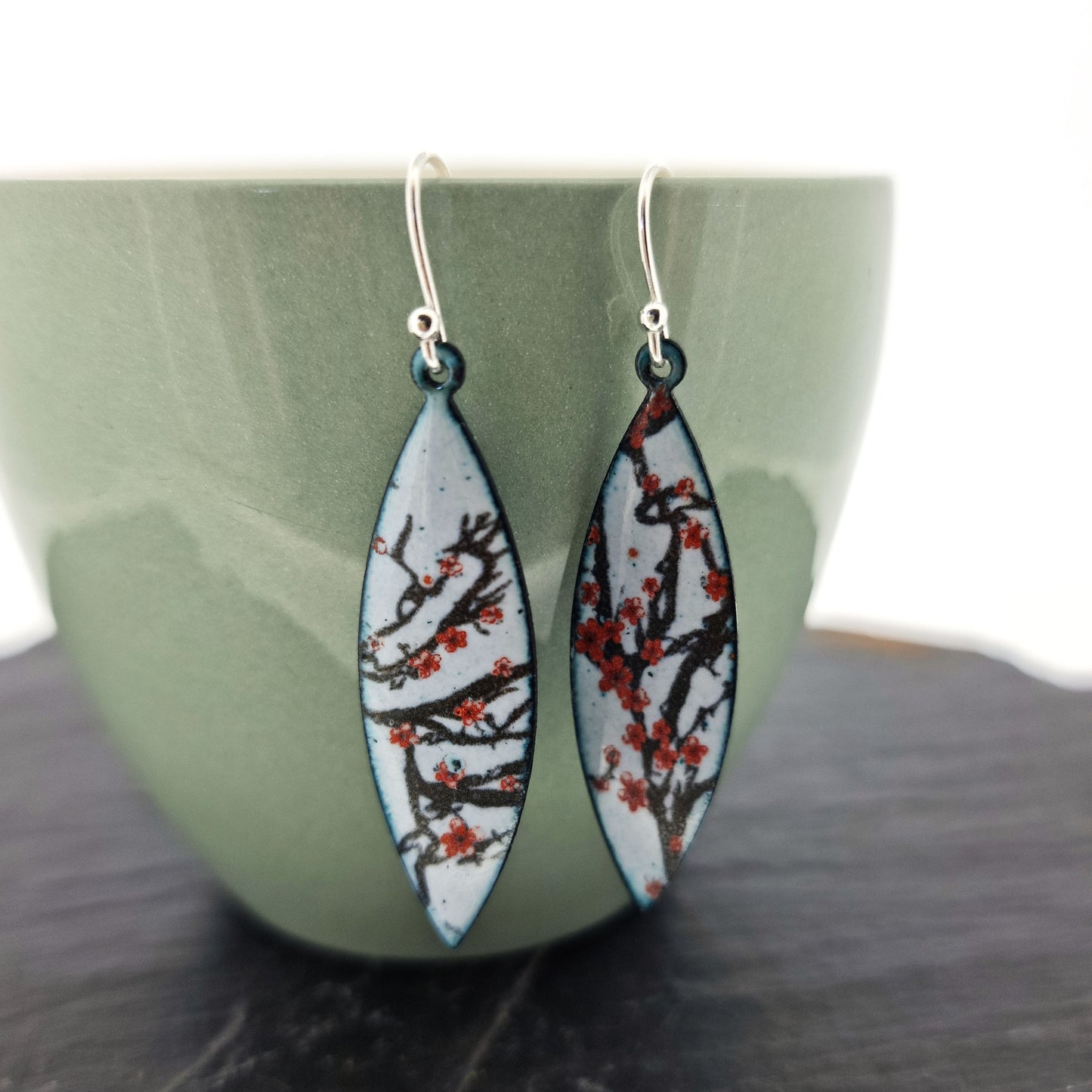 Boat-shaped enamel drop earrings with branches of red cherry blossom on a grey background. Pictured on a cup.