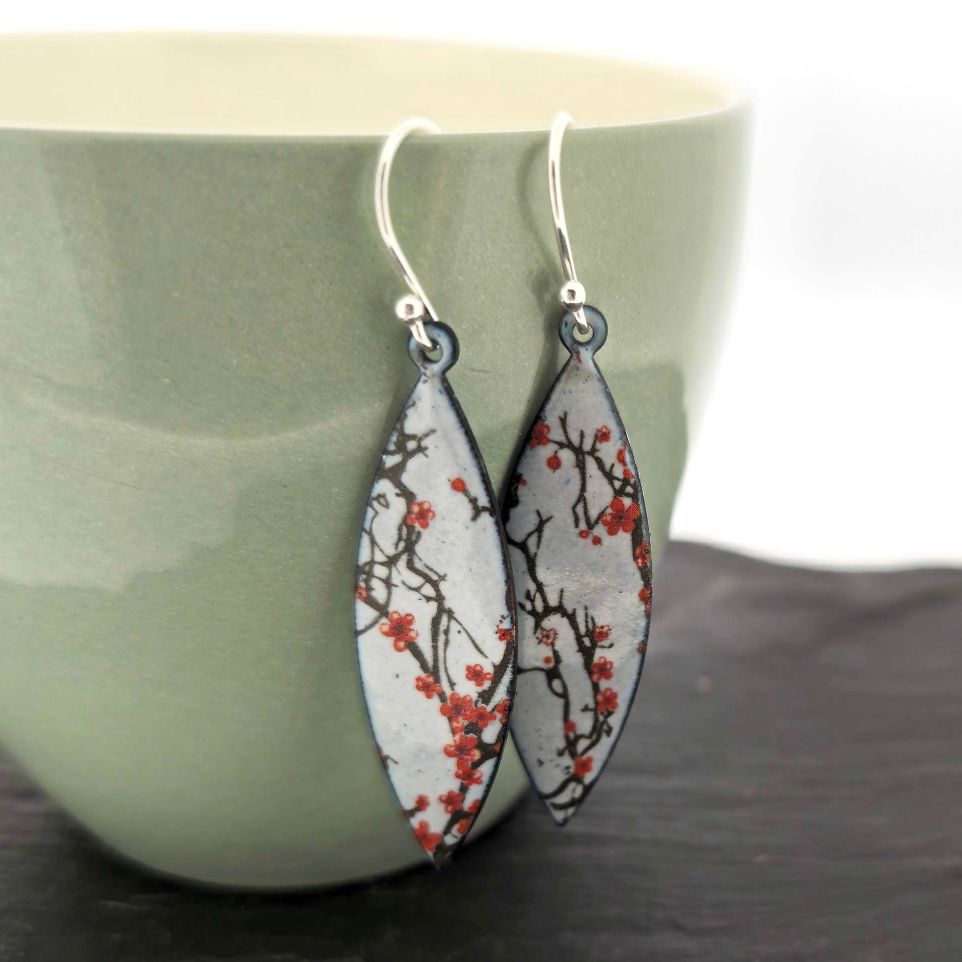 Pair of enamel drop earrings with cherry blossom design on a green ceramic cup