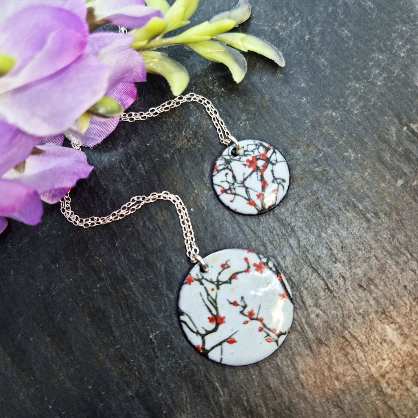 Two round enamel pendants each on a silver chain. The pendants have a light grey background with branches running across it and red blossom flowers. Pictured on slate with flowers.