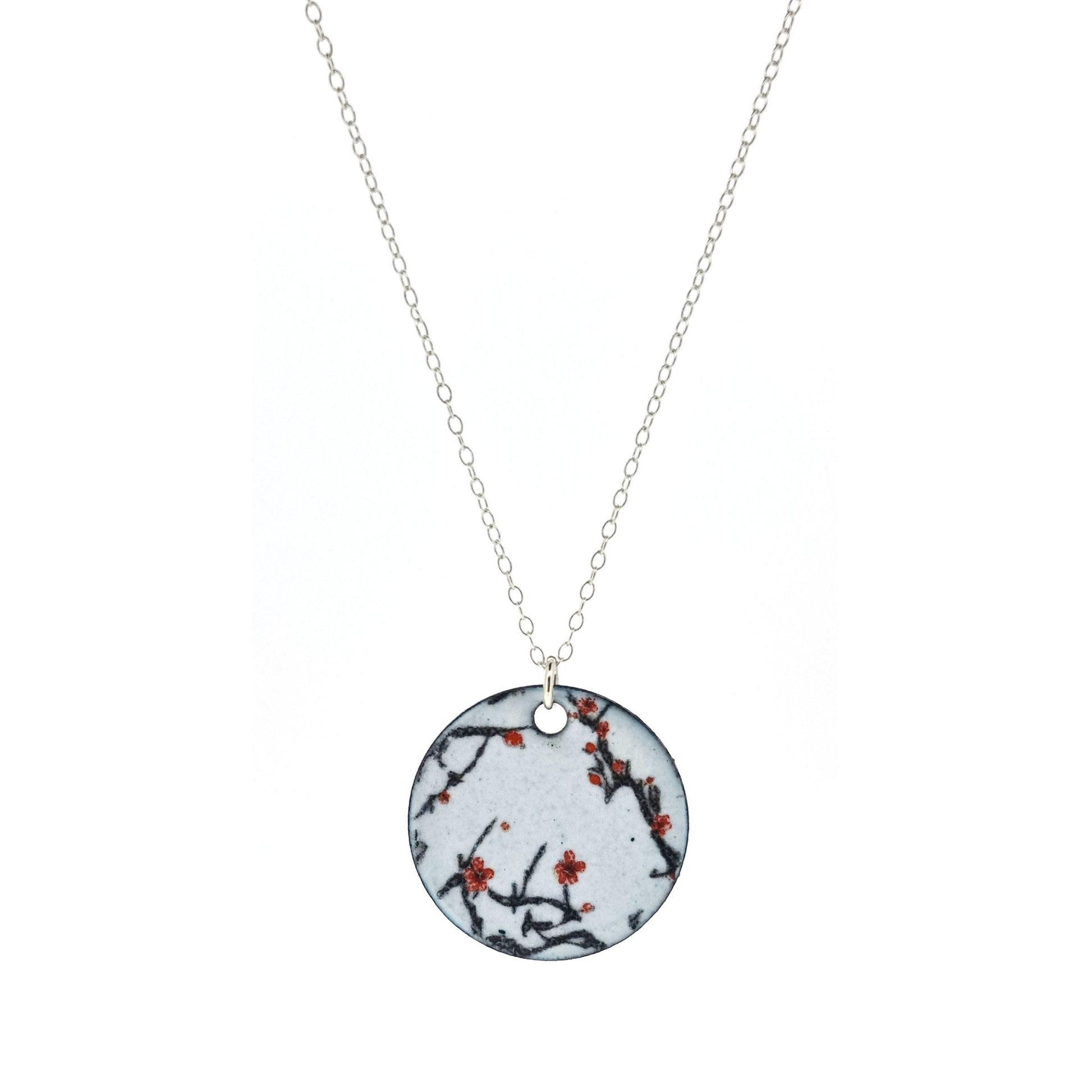 A round enamel pendant hangs from a silver chain. The pendant has a light grey background with branches running across it and red blossom flowers.