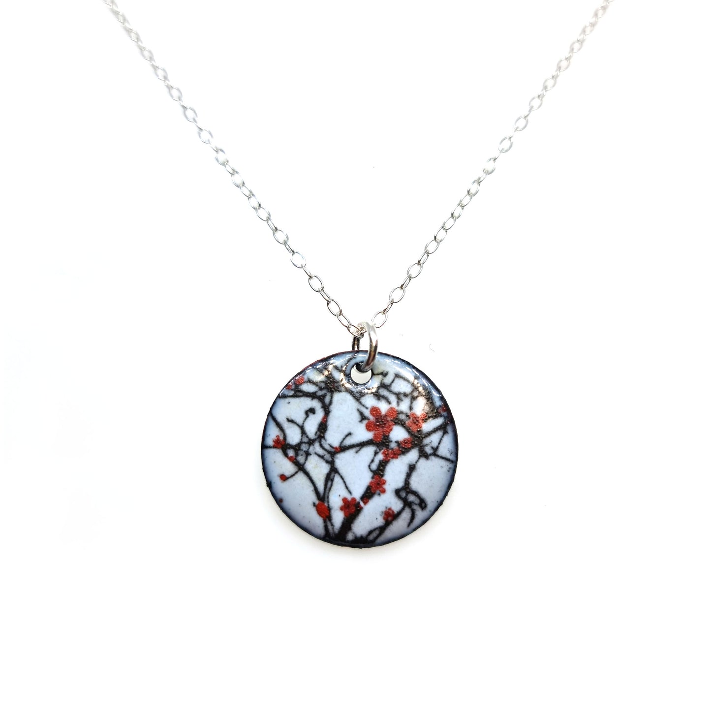 A round enamel pendant hangs from a silver chain. The pendant has a light grey background with branches running across it and red blossom flowers.