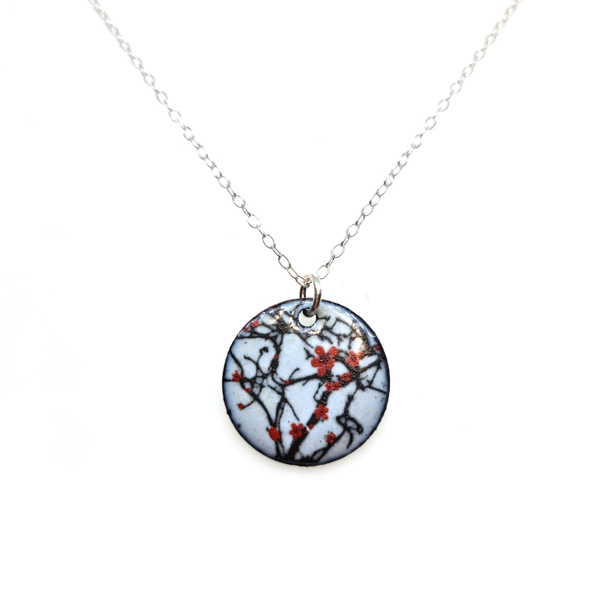 A round enamel pendant hangs from a silver chain. The pendant has a light grey background with branches running across it and red blossom flowers.