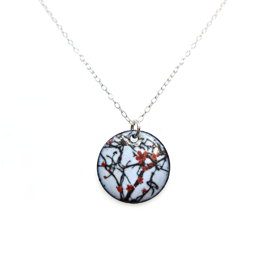A round enamel pendant hangs from a silver chain. The pendant has a light grey background with branches running across it and red blossom flowers.