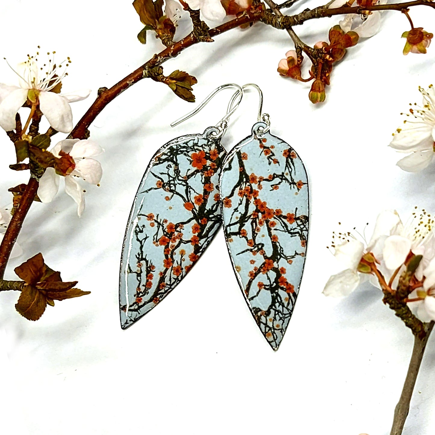 Statement enamel earrings featuring branches and red cherry blossom. On silver ear hooks. Surrounded by white cherry blossom branches.