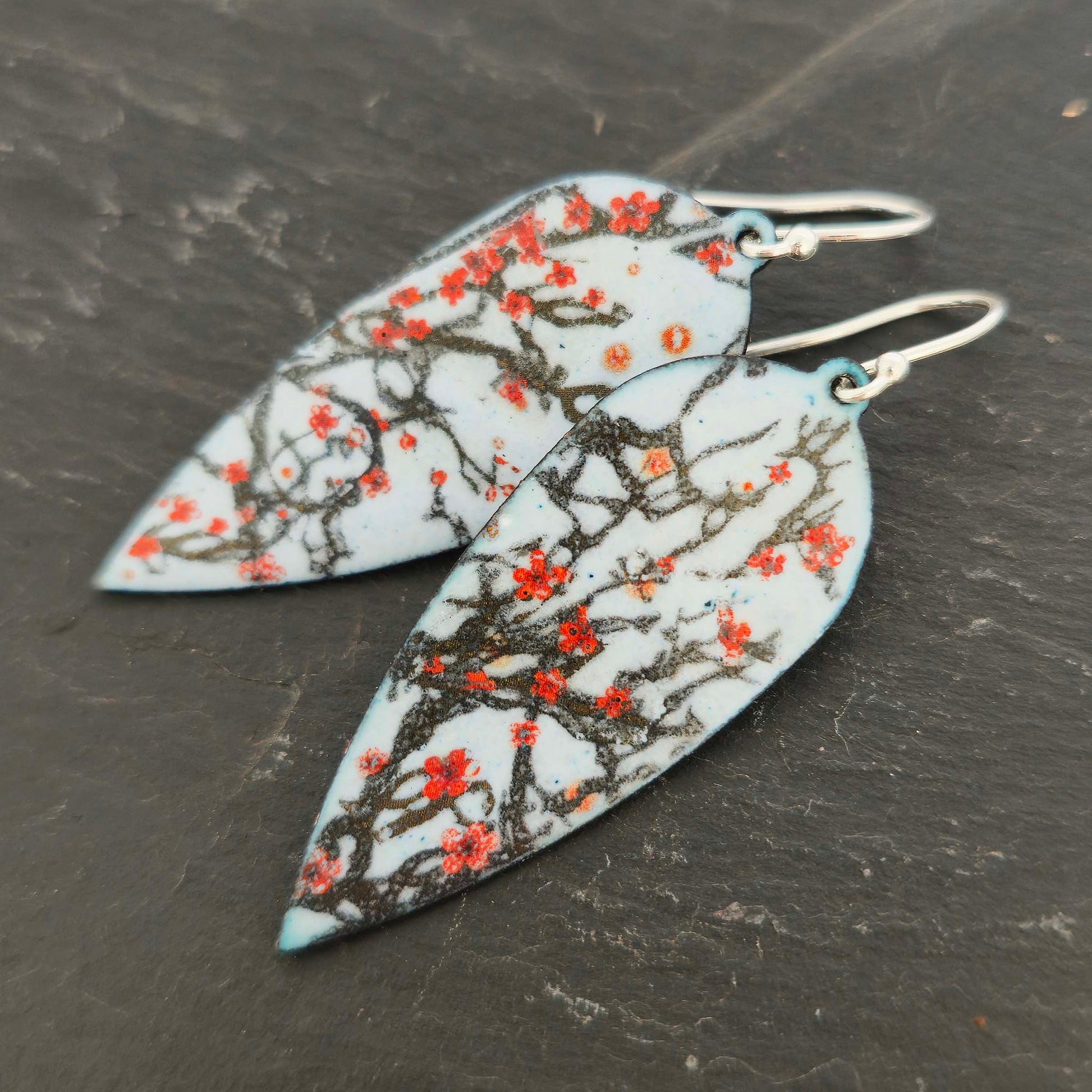 Statement enamel earrings featuring branches and red cherry blossom on a grey enamel background. On silver ear hooks. 