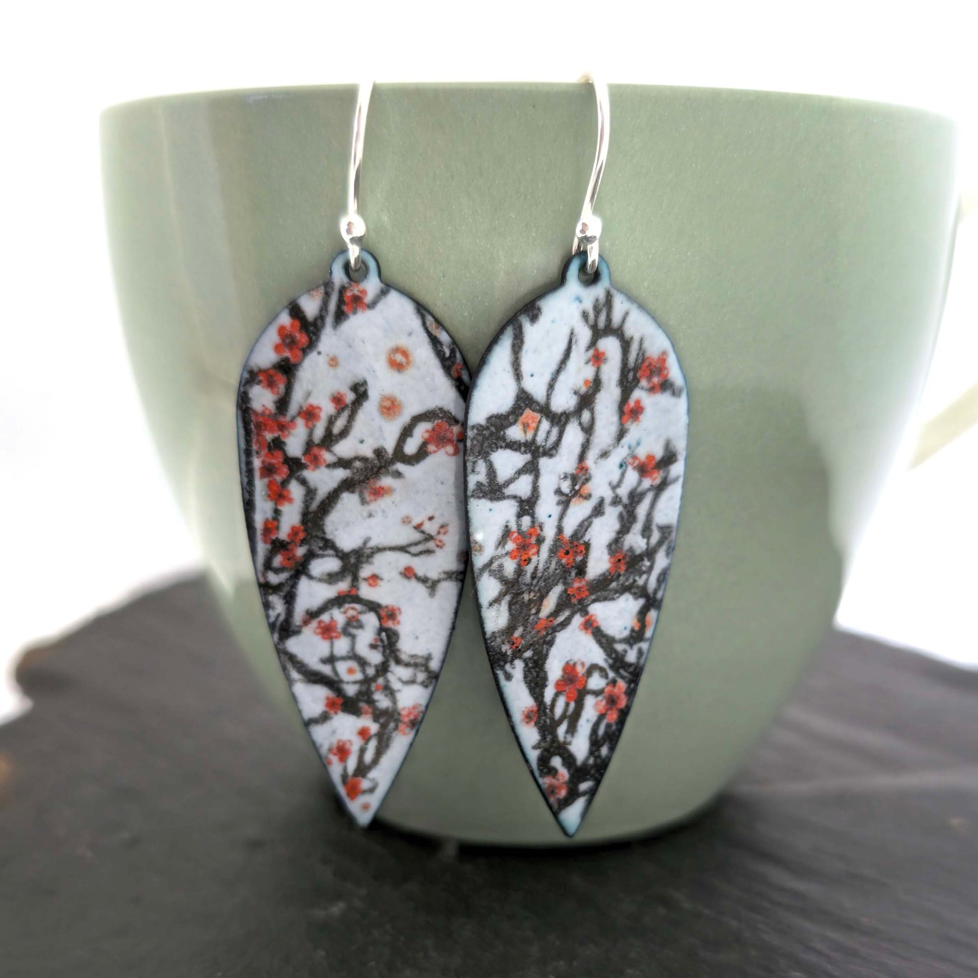 Statement enamel earrings featuring branches and red cherry blossom on a grey enamel background. On silver ear hooks. Shown on a green cup.