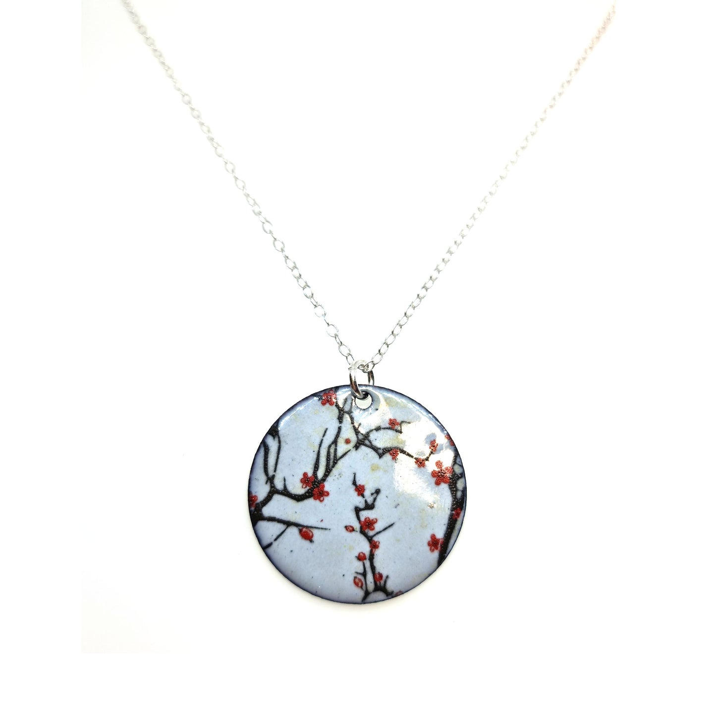 A round enamel pendant hangs from a silver chain. The pendant has a light grey background with branches running across it and red blossom flowers.