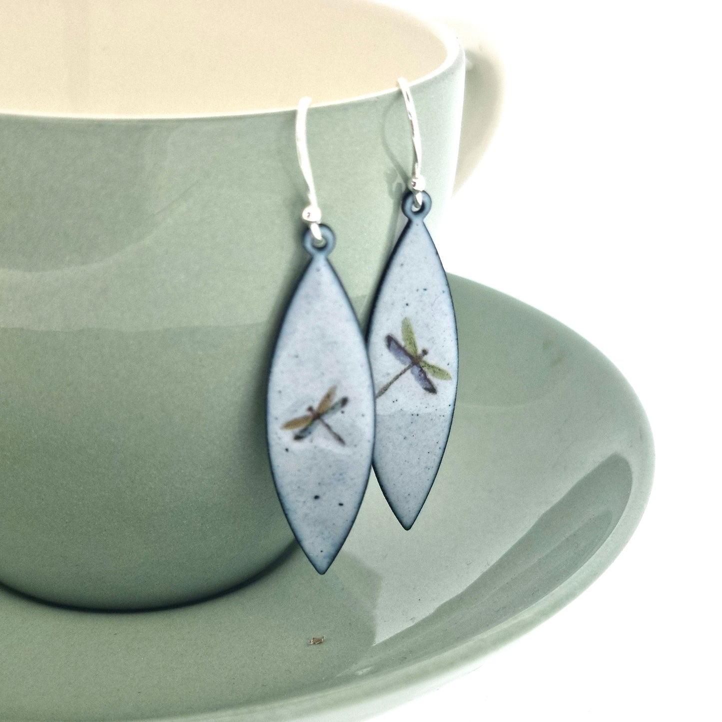 Boat shaped drop earrings with grey enamel. Each earring features one dragonfly in various colours. On silver ear hooks. Pictured on a cup.