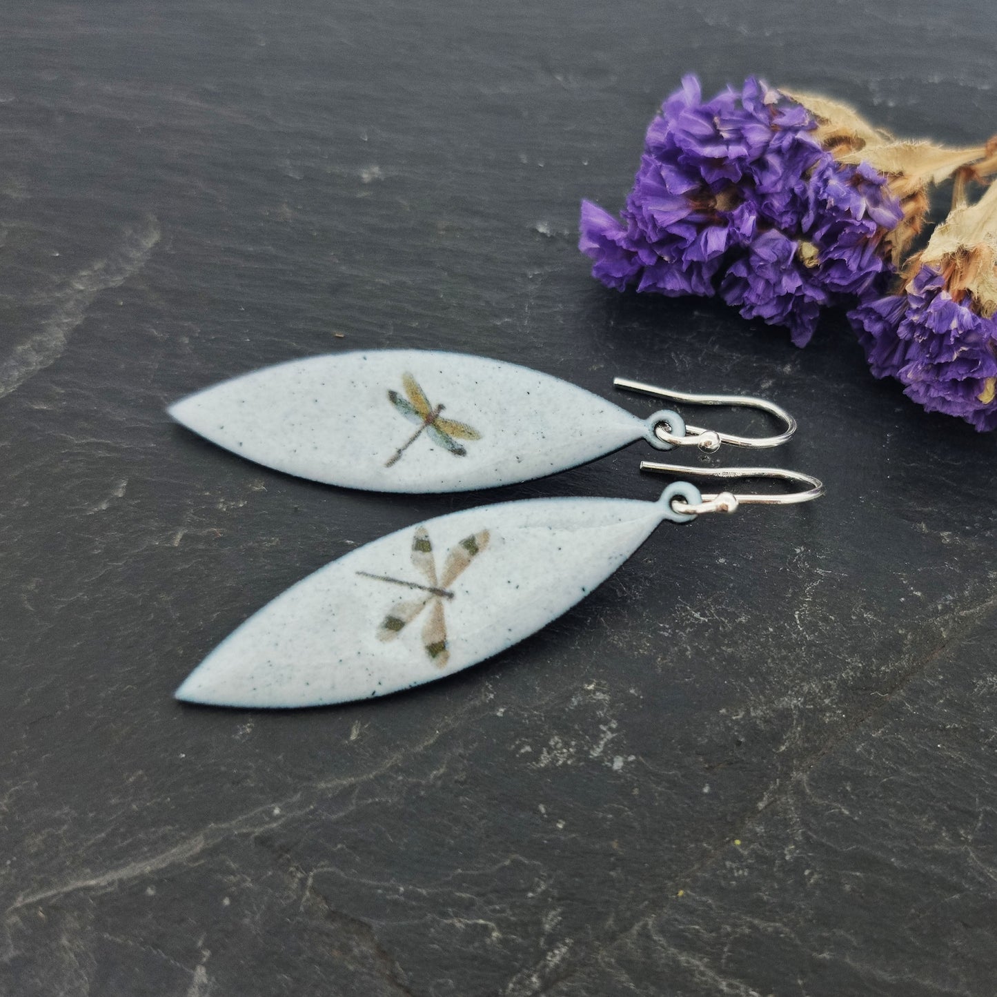 Boat shaped drop earrings with grey enamel. Each earring features one dragonfly in various colours. On silver ear hooks. Pictured on slate with flowers.