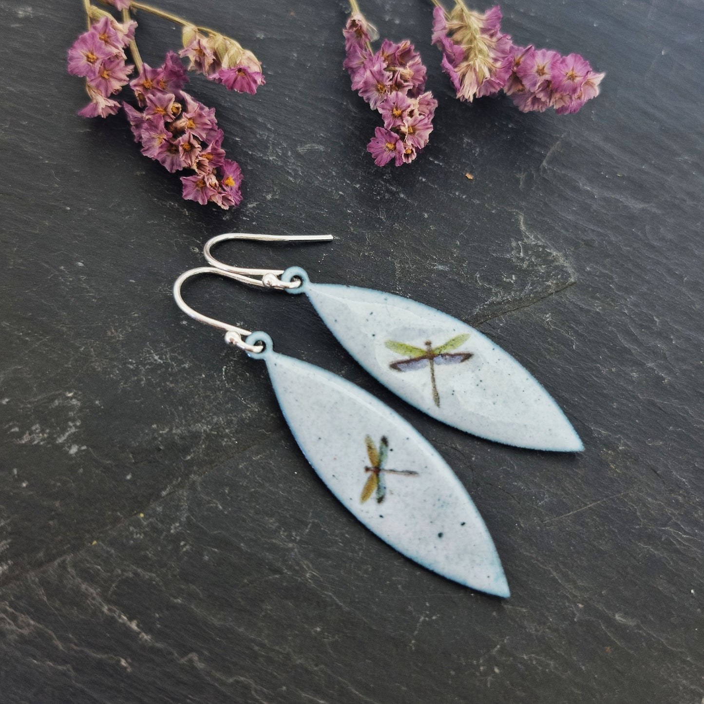 Boat shaped drop earrings with grey enamel. Each earring features one dragonfly in various colours. On silver ear hooks. Pictured on slate with flowers.