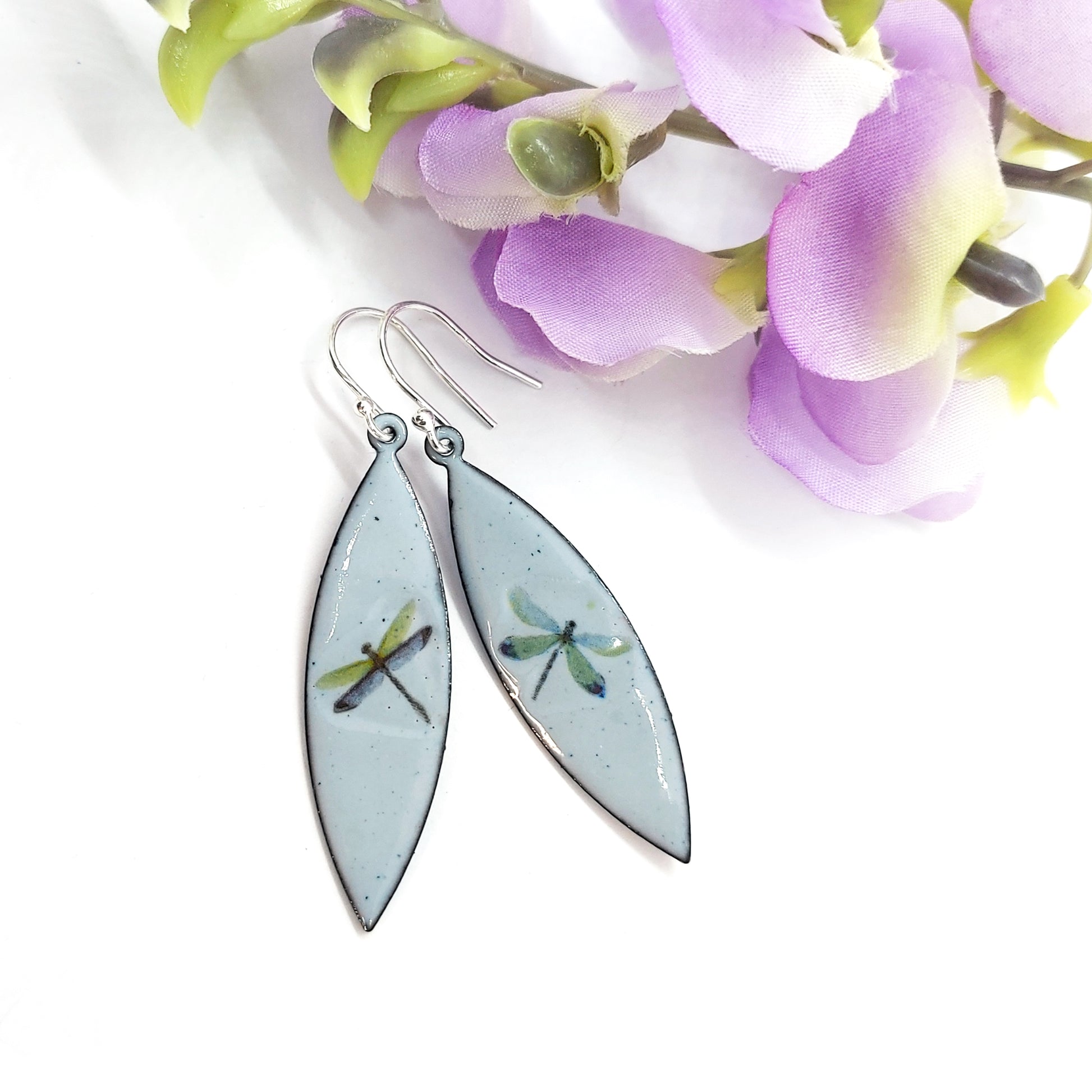 Boat shaped drop earrings with grey enamel. Each earring features one dragonfly in various colours. On silver ear hooks. Pictured with flowers.