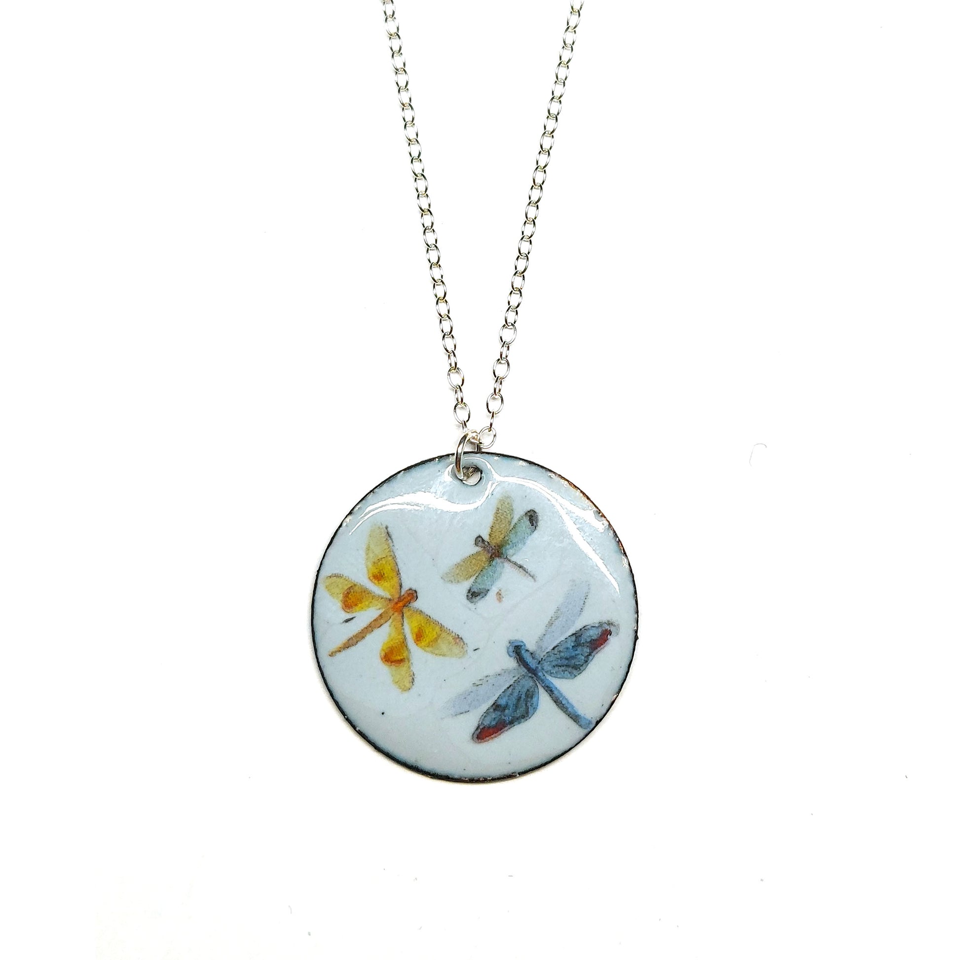 A round enamel pendant hangs from a silver chain. The pendant has a light grey background and 3 dragonflies on it.