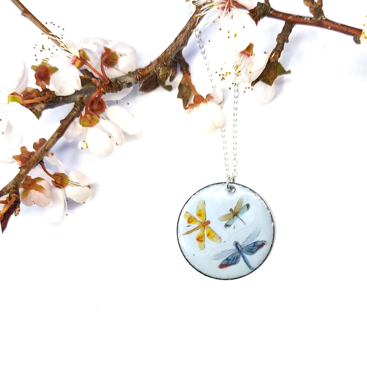 A round enamel pendant hangs from a silver chain. The pendant has a light grey background and 3 dragonflies on it - with branch