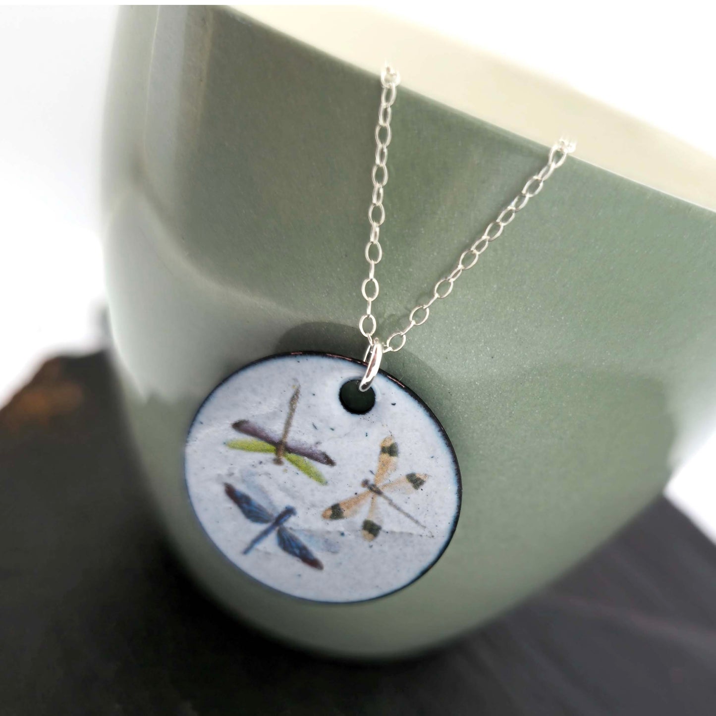 Necklace with a round enamel pendant featuring three dragonfly designs on a silver chain. Shown on a ceramic cup.