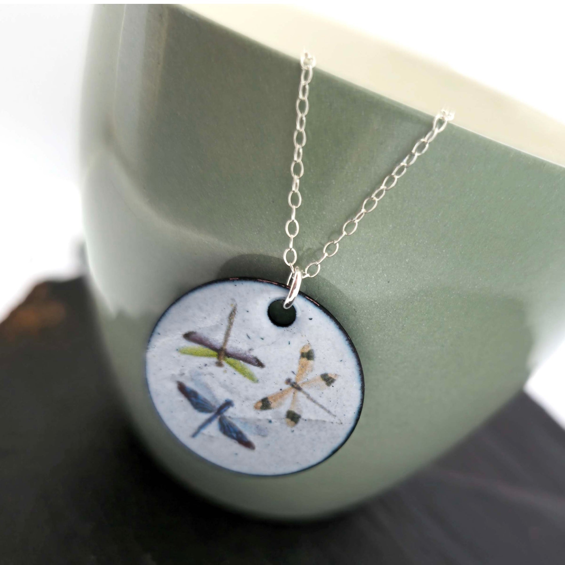 Necklace with a round enamel pendant featuring three dragonfly designs on a silver chain. Shown on a ceramic cup.