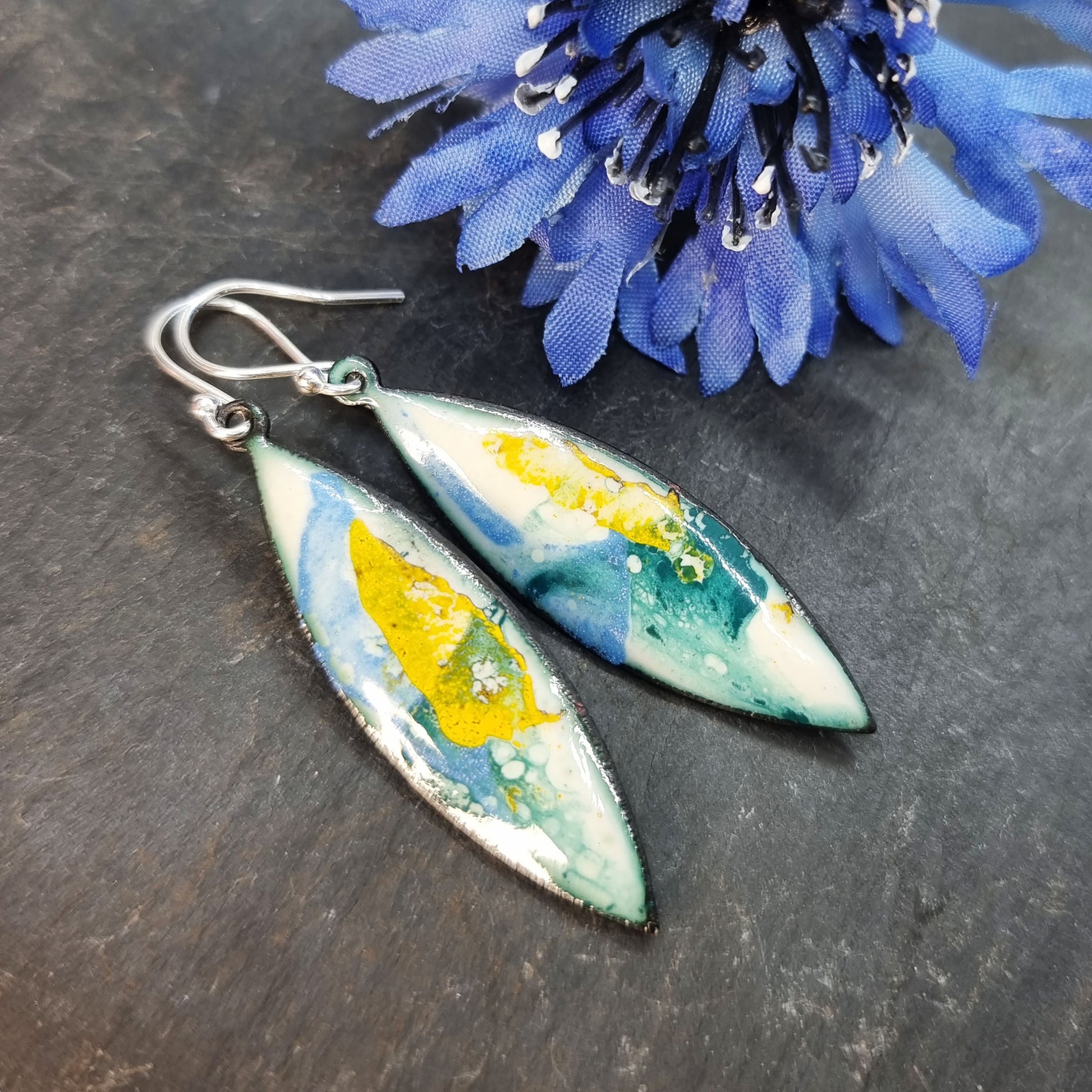 Navette shaped enamel drop earrings with green, blue and yellow splashes on a white background - with flower