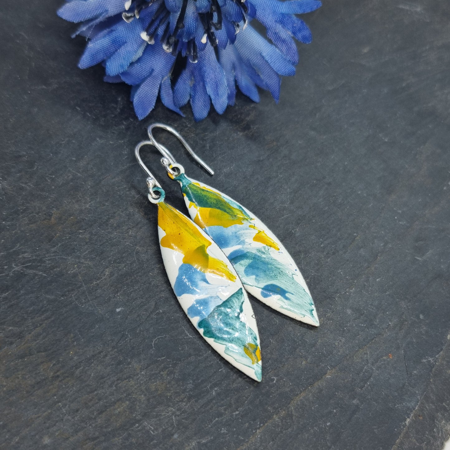 Navette shaped enamel drop earrings with green, blue and yellow splashes on a white background - with flower