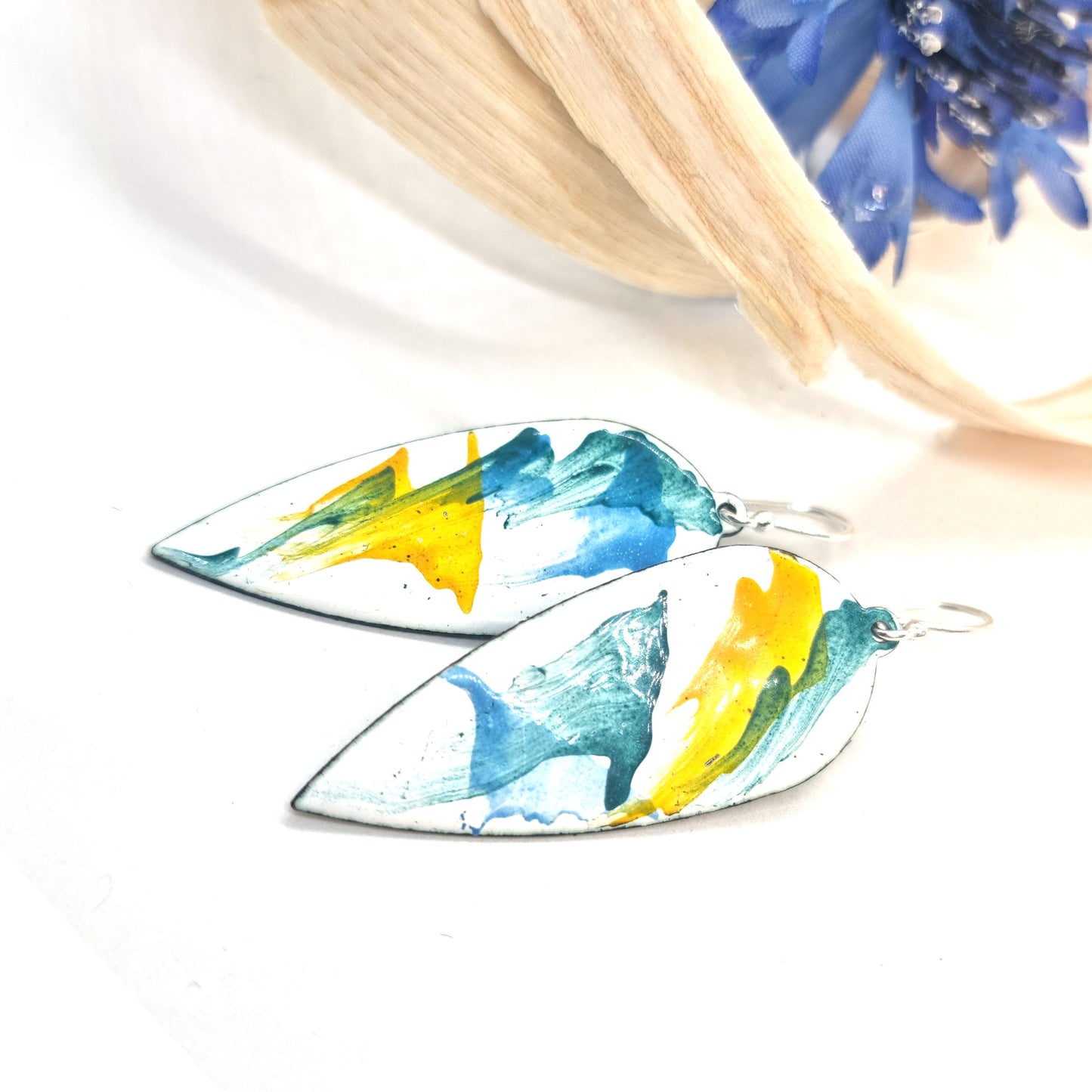 Large teardrop-shaped enamel drop earrings with splashes of green, blue and yellow on a white background - with flower and wood shavings