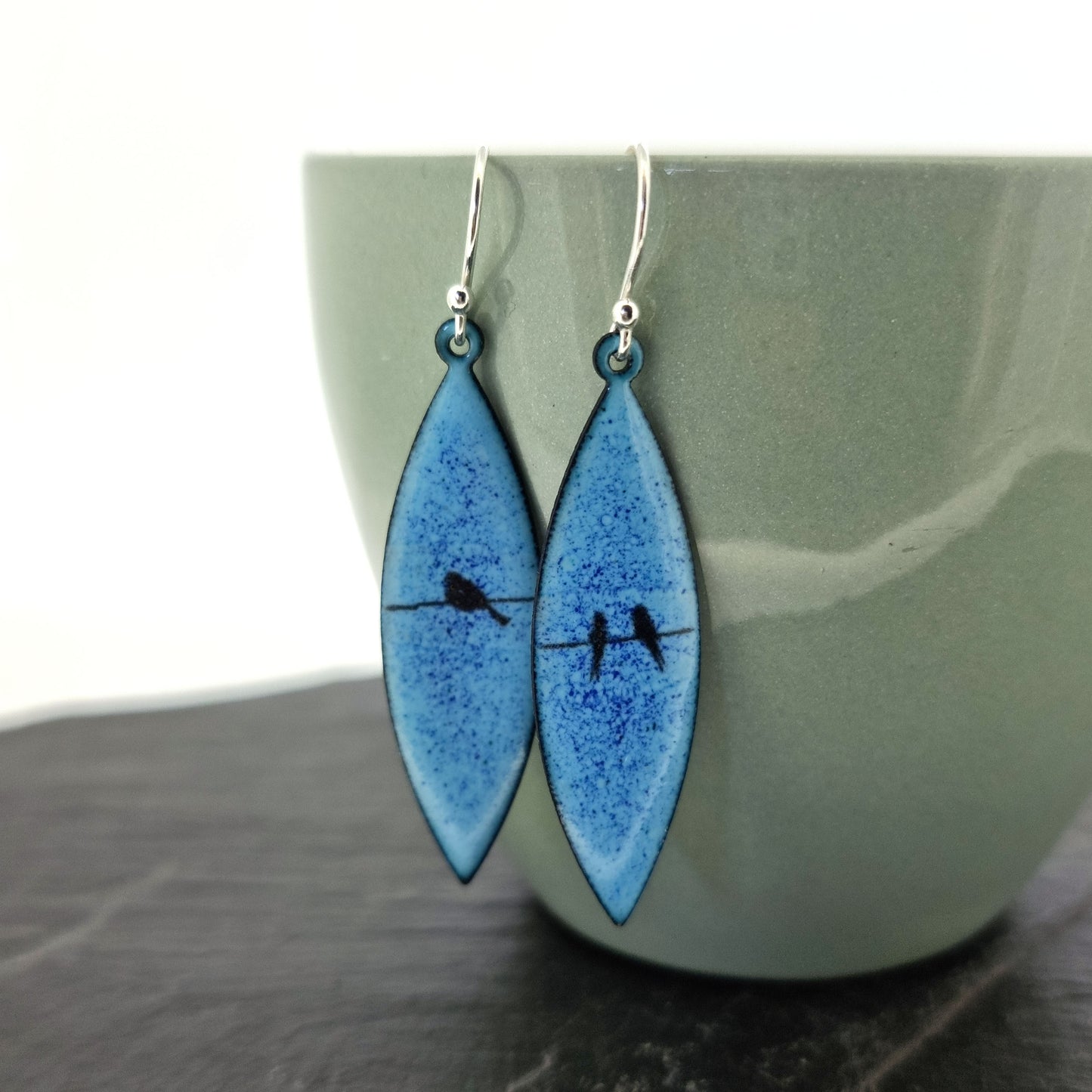 Boat-shaped enamel drop earrings with black bird silhouettes sitting on a wire on a blue background - pictured on a cup.