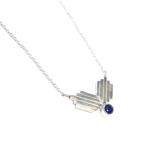 A silver Art Deco style necklace with a focal point of 2 lined elements and a dark blue sapphire gemstone.