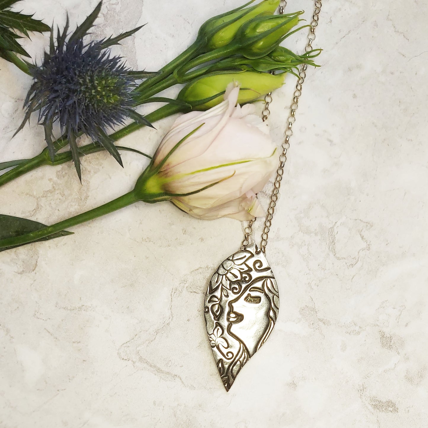 Silver leaf-shaped pendant on silver chain. The pendant features a smiling woman and a flower - with flowers