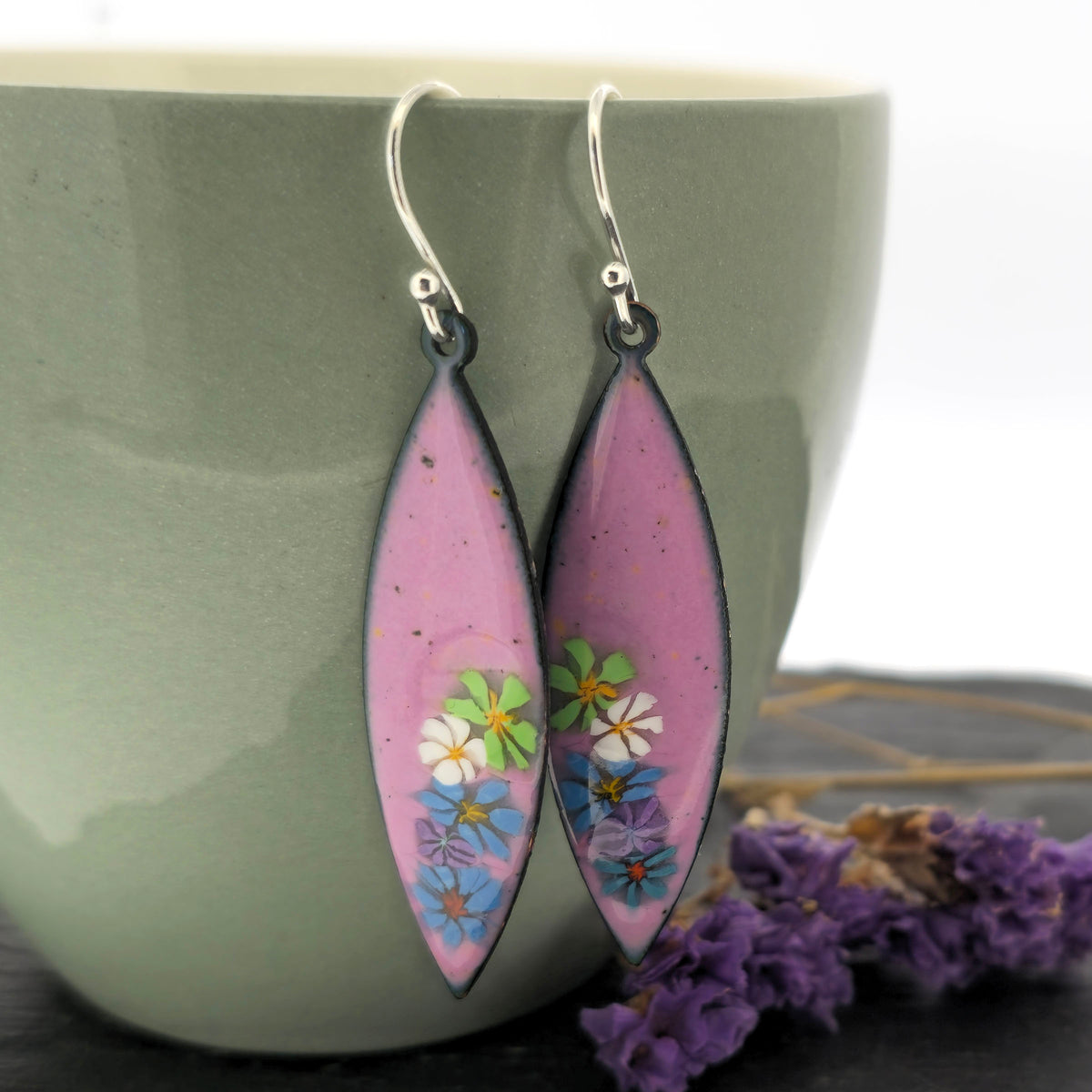 A pair of handmade marquise shaped drop earrings with a background of pink enamel and colourful enamel flowers along one edge. On silver ear hooks. Shown on a sage green cup. Mijoux jewellery.