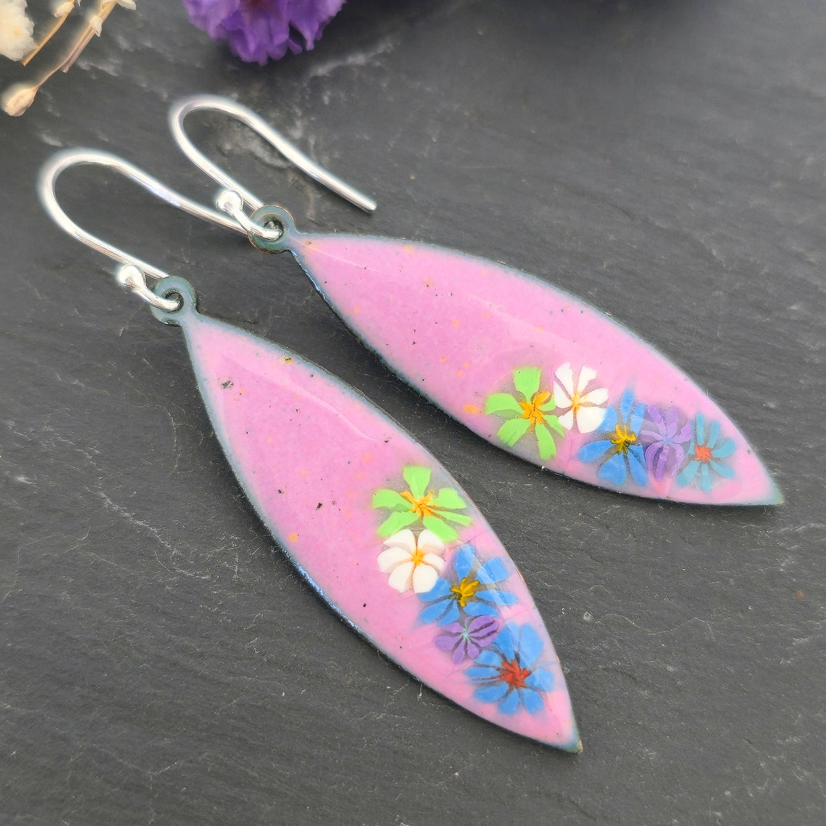 A pair of handmade marquise shaped drop earrings with a background of pink enamel and colourful enamel flowers along one edge. On silver ear hooks. Shown on a black background. Mijoux jewellery.