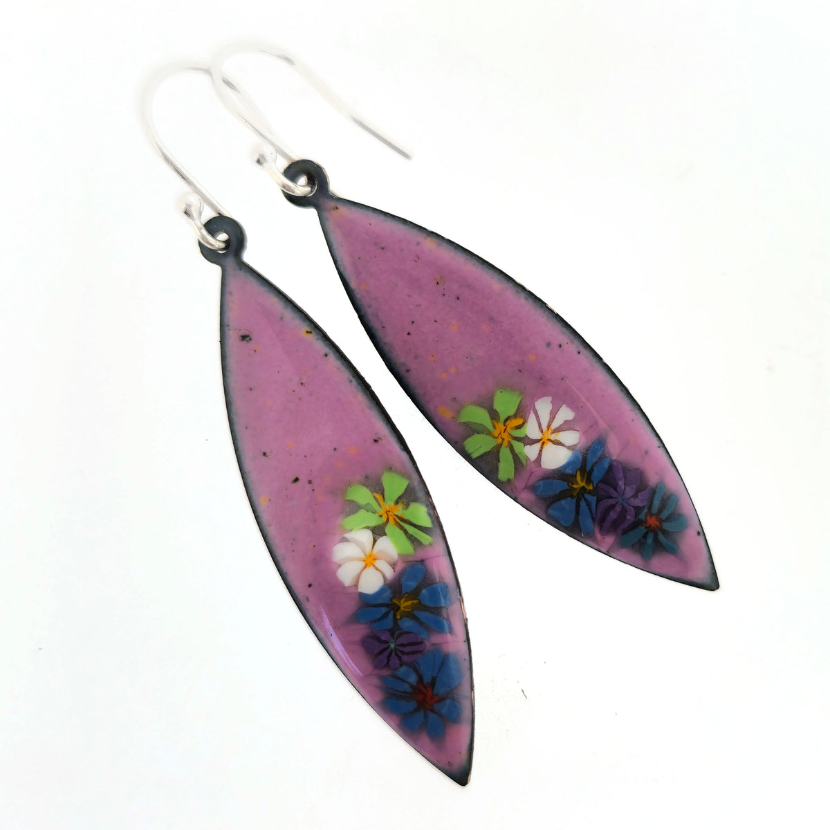 A pair of handmade marquise shaped drop earrings with a background of pink enamel and colourful enamel flowers along one edge. On silver ear hooks. Shown on a white background. Mijoux jewellery.