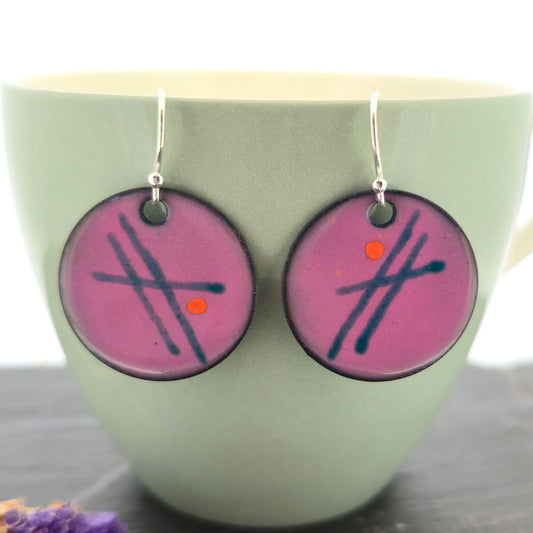 A pair of handmade round pink enamel drop earrings with green lines and orange dots. On silver ear hooks. On a pale green cup. Mijoux jewellery.