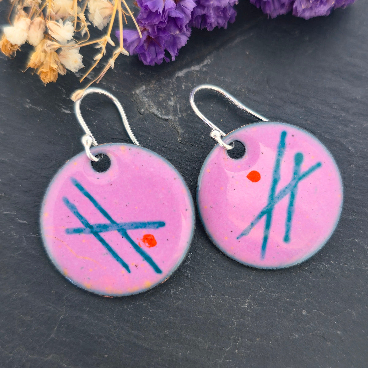 A pair of handmade round pink enamel drop earrings with green lines and orange dots. On silver ear hooks. On a black background with purple flowers. Mijoux jewellery.