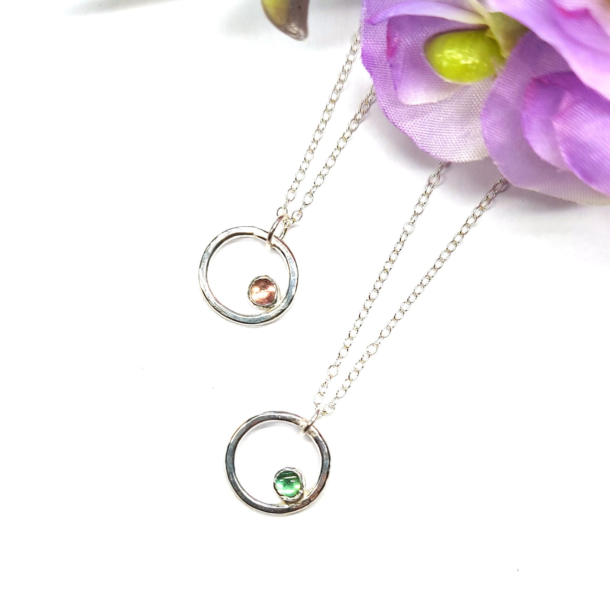 Silver hammered open circle pendants with round green or pink tourmaline gemstone set off-centre. On a silver chain. With flowers.