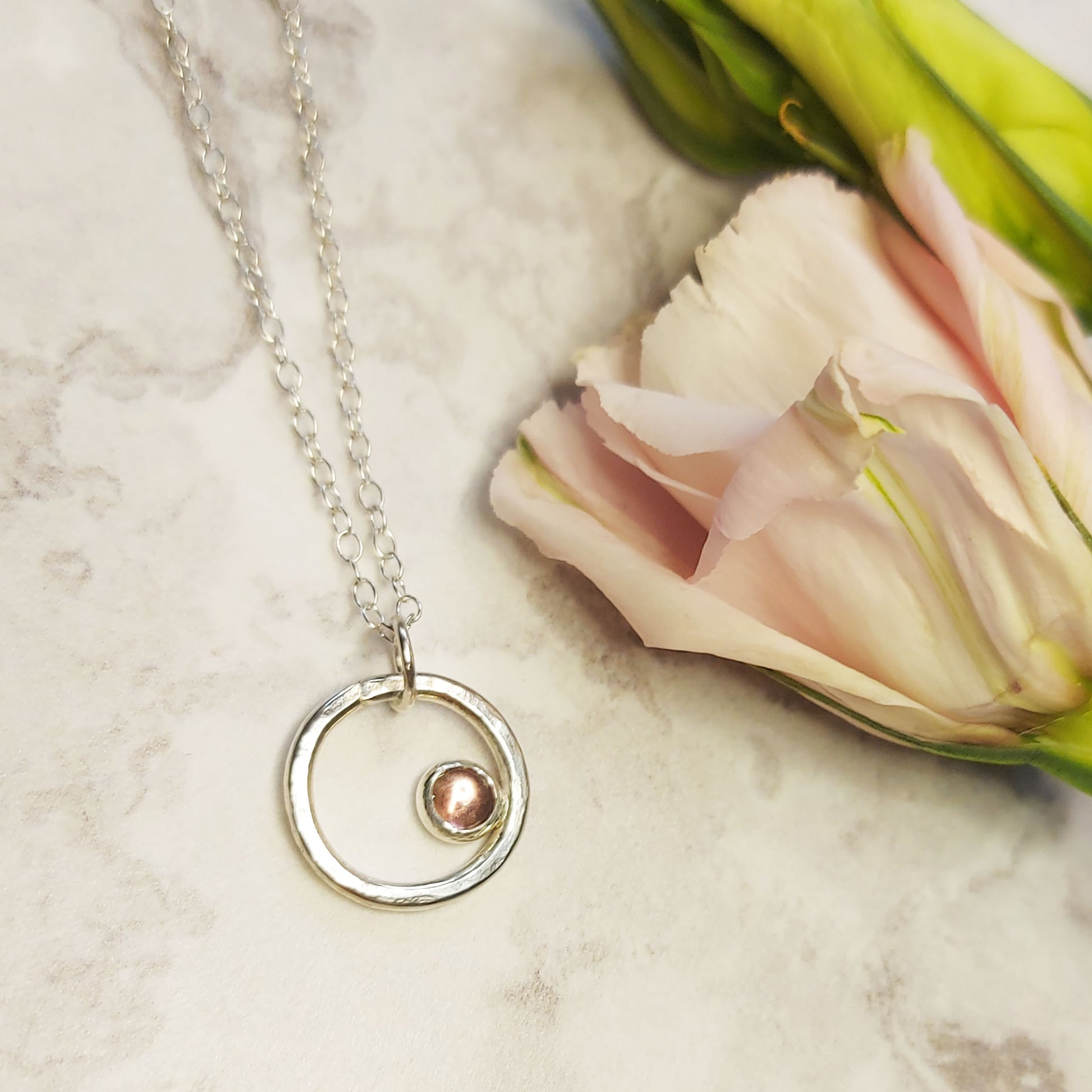 Silver hammered open circle pendant with round pink tourmaline gemstone set off-centre. On a silver chain. With flowers.