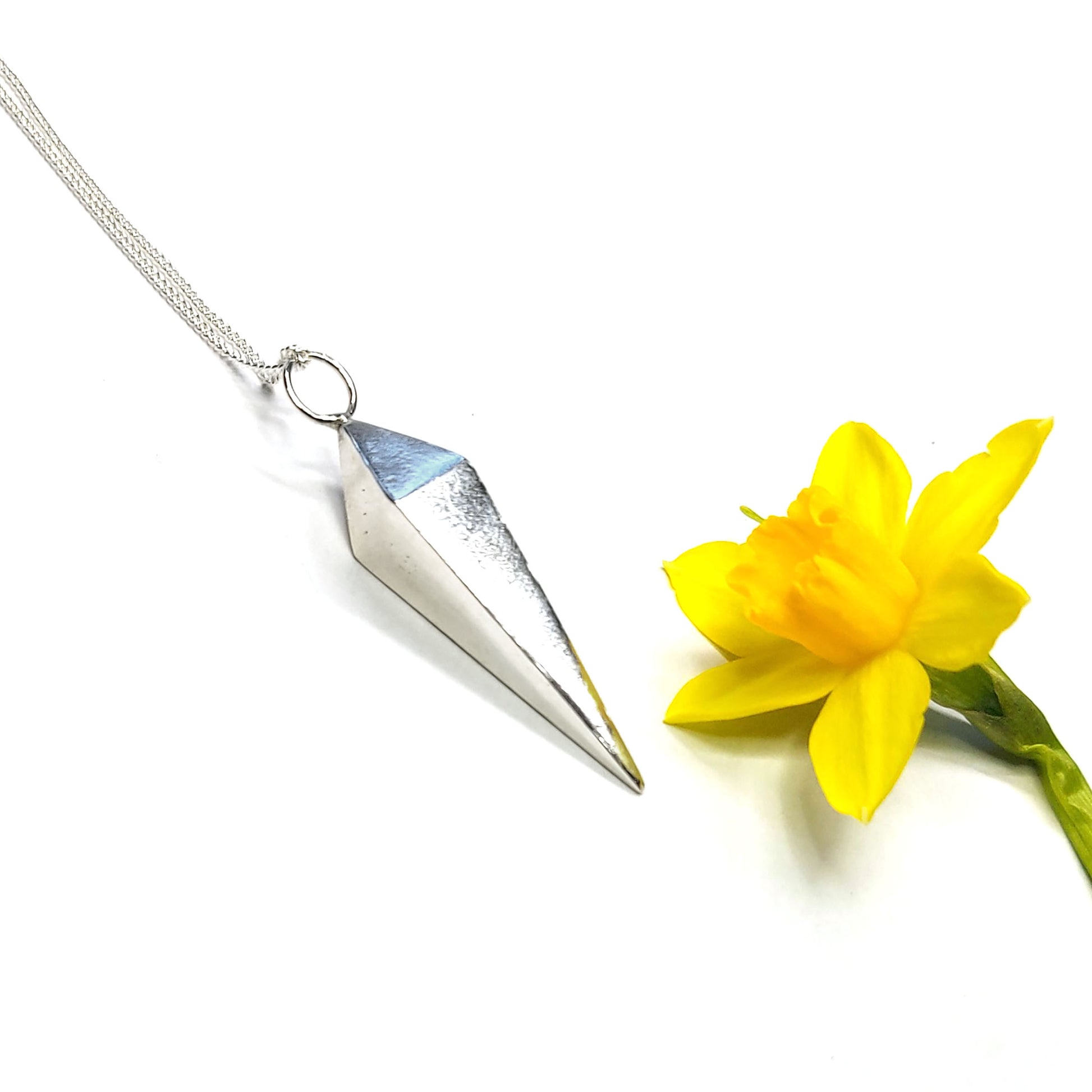 A silver geometric diamond shaped pendant on a silver chain. Pictured with a daffodil.