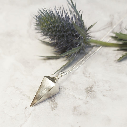 A silver 3D diamond shaped pendant on a silver chain. Pictured with a thistle flower.