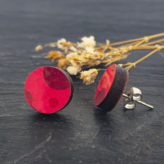 A pair of wooden stud earrings with a vibrant red circular pattern and natural brown wood edges, displayed on a dark textured surface with dried flowers in the background. Mijoux Jewellery.