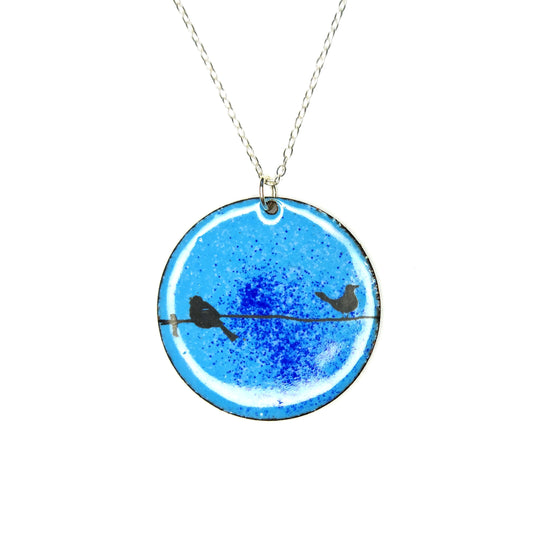 Round enamel pendant with bird silhouettes sat on a wire against a blue background. Suspended on a silver chain.