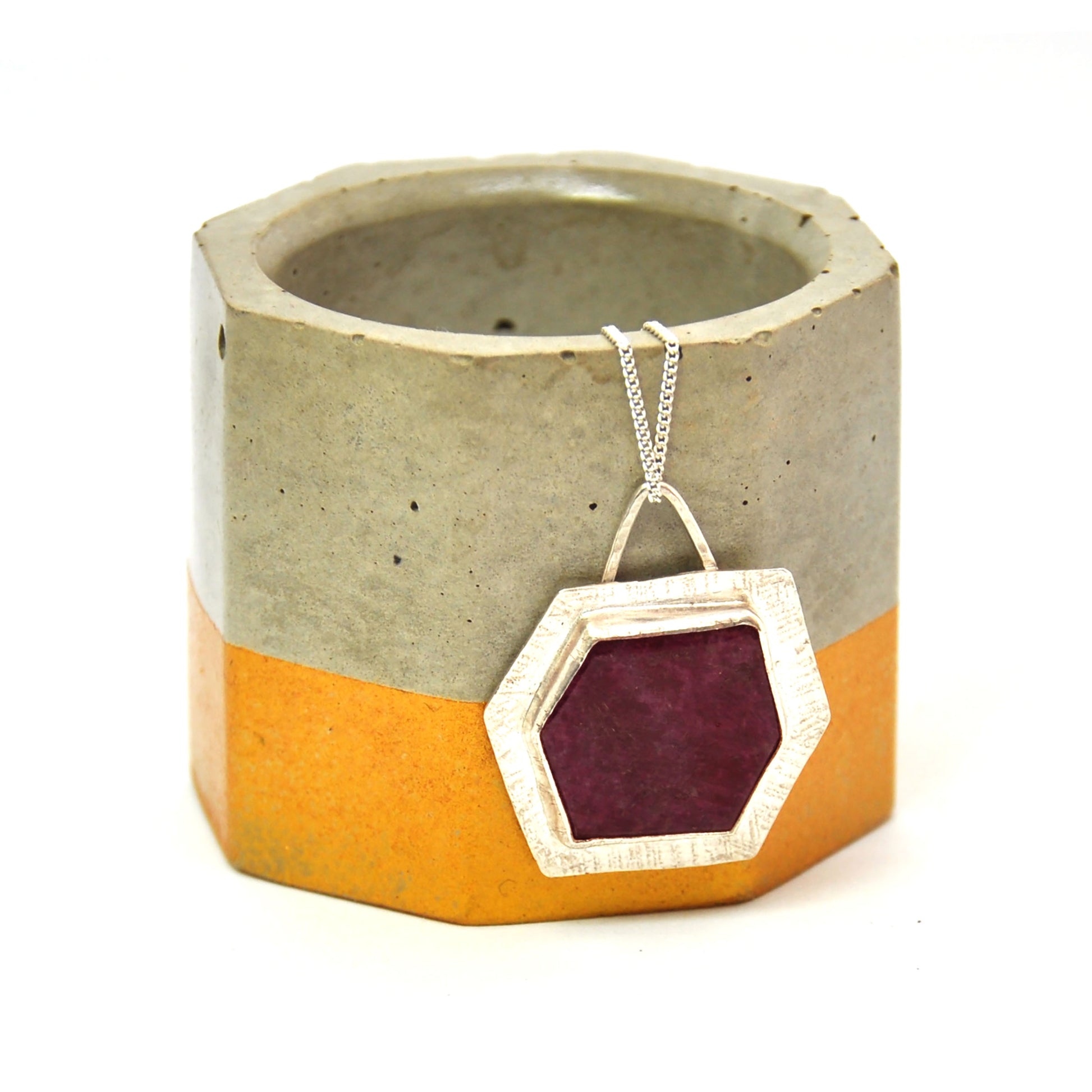 A silver pendant with a hexagonal ruby slice set into a textured surround. On a silver chain. Pictured on a vase.