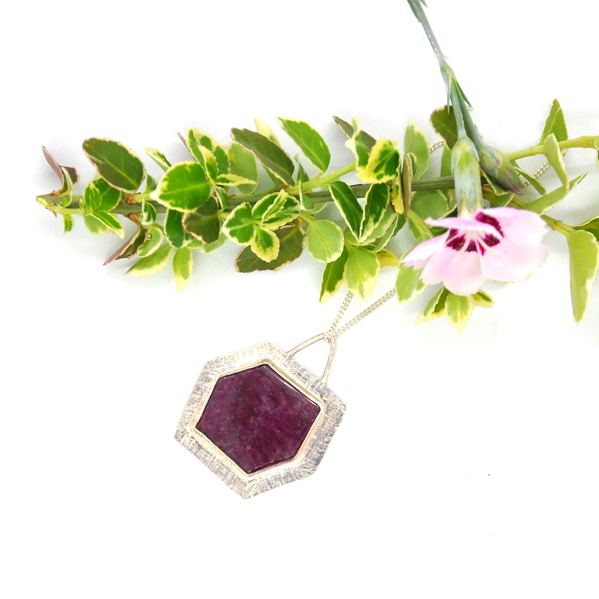 A silver pendant with a hexagonal ruby slice set into a textured surround. On a silver chain. Pictured with flowers.