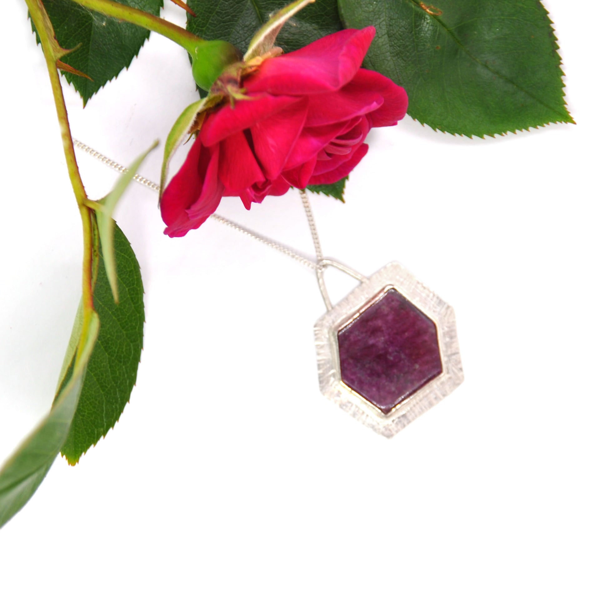 A silver pendant with a hexagonal ruby slice set into a textured surround. On a silver chain. Pictured with a rose.